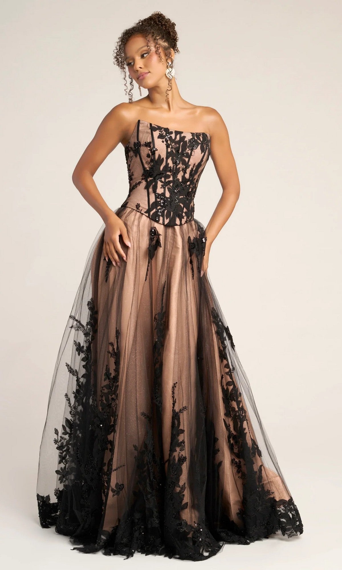 A woman wears the Portia and Scarlett PS26061 long formal dress in black/nude, featuring an a-line silhouette, strapless corset bodice, floral lace overlay, nude underlayer, and a full glitter tulle skirt.